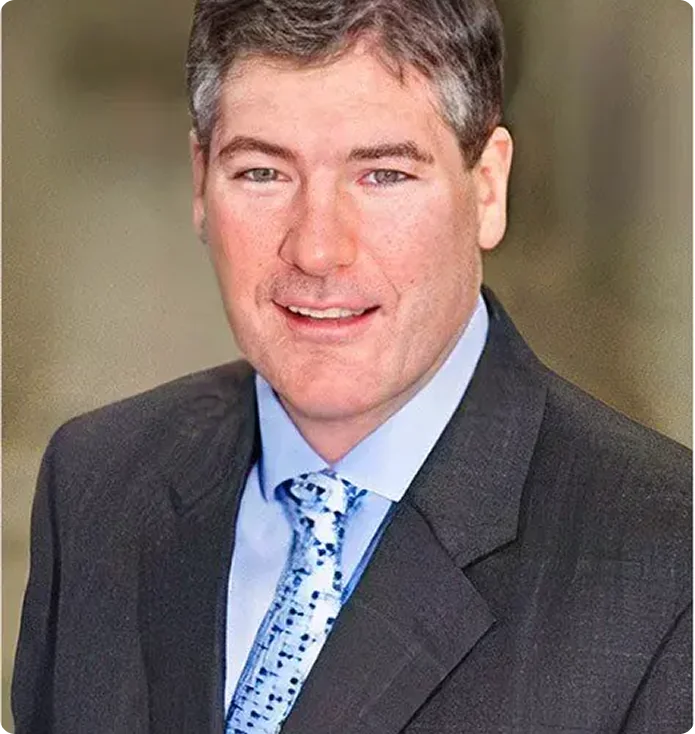 A man with short brown hair is wearing a dark suit, light blue dress shirt, and a patterned blue tie, standing in front of a neutral, blurred background.